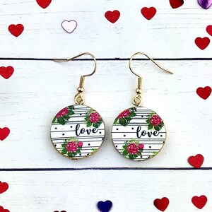 Może przedstawiać: Para złotych kolczyków wiszących z okrągłymi, czarno-białymi, paskowanymi drewnianymi dyskami. Dyski mają kwiatowy wzór z różowymi różami i nadrukowanym słowem "love".