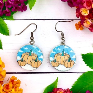 Può includere: Un paio di orecchini d'argento con un design rotondo blu e bianco con tre zucche arancioni. Le zucche sono impilate l'una sull'altra e hanno steli verdi.