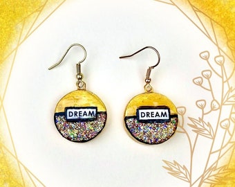 Può includere: Un paio di orecchini pendenti circolari con una metà superiore color oro e una metà inferiore con glitter multicolore. Un'etichetta rettangolare bianca con il testo nero DREAM è centrata su ogni orecchino. Pendono da ganci in metallo dorato.