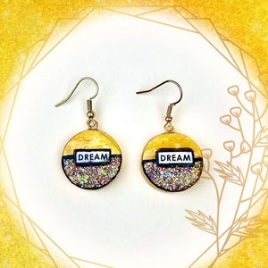 May include: A pair of circular drop earrings featuring a gold-colored top half and a multi-colored glitter bottom half. A rectangular white label with the black text DREAM is centered on each earring. The earrings hang from gold-toned metal hooks.