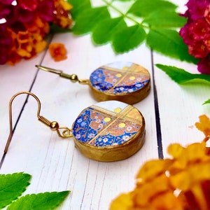 May include: A pair of gold earrings with blue, white, and gold patterned round ceramic charms. The charms have a floral design with gold accents.