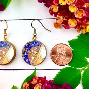 May include: A pair of gold dangle earrings with blue and white porcelain round pendants. The pendants have a floral pattern and gold accents. The earrings are shown next to a US penny for scale.