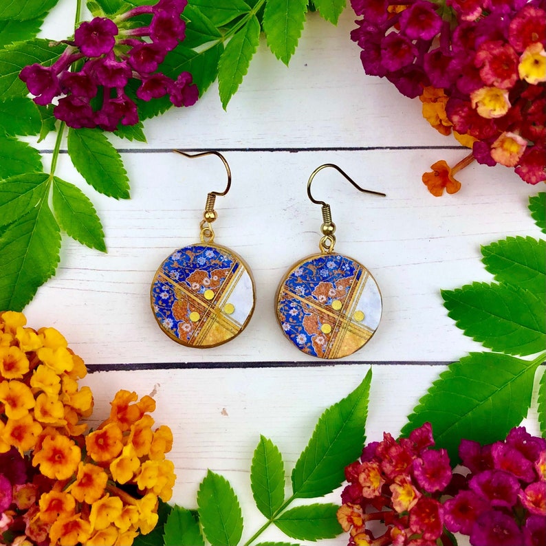 May include: A pair of gold dangle earrings with round blue, white, and gold patterned ceramic charms. The charms have a floral design with gold accents.