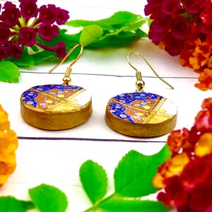 May include: A pair of gold-toned earrings with round blue, white, and gold patterned ceramic pendants. The pendants have a floral design with gold accents.