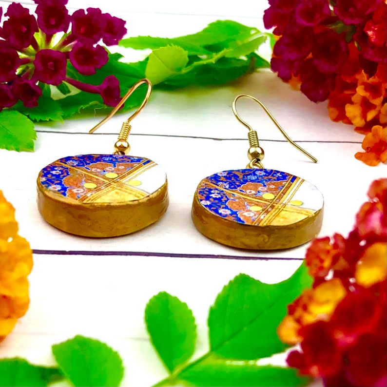 May include: A pair of gold-toned earrings with round blue, white, and gold patterned ceramic pendants. The pendants have a floral design with gold accents.