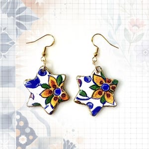 May include: A pair of star-shaped dangle earrings featuring a white base with a hand-painted floral pattern in shades of yellow, orange, green, and blue. The earrings are suspended from gold-toned metal fishhook wires and have a glossy finish.