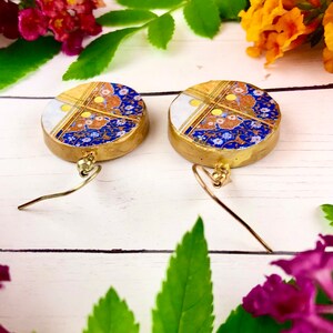May include: A pair of gold dangle earrings with round blue, white, and gold patterned ceramic charms. The charms have a floral design with gold accents.