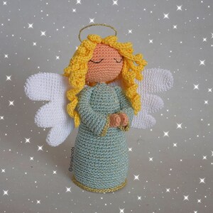 May include: A crocheted angel figurine with a blue dress, yellow hair, and white wings. The angel has a halo and is holding its hands together in prayer.