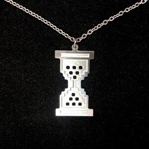 Könnte beinhalten: Eine silberfarbene Halskette mit einem pixeligen Sanduhr-Anhänger. Der Anhänger hat ein geometrisches Design mit kleinen quadratischen Aussparungen. Die Halskette ist zierlich und silberfarben, vor schwarzem Hintergrund.