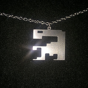 Puede incluir: Un collar de cadena de plata con un colgante de plata con forma de dinosaurio pixelado.