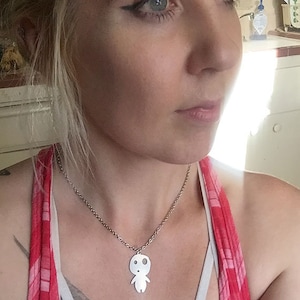 May include: A silver-colored pendant necklace featuring a stylized figure with a round head and small arms and legs. The pendant hangs from a delicate chain. The person is wearing a red and pink plaid top.