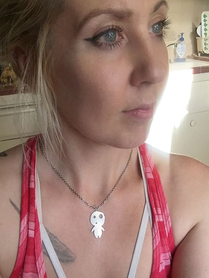 May include: A silver-colored pendant necklace featuring a stylized figure with a round head and small arms and legs. The pendant hangs from a delicate chain. The person is wearing a red and pink plaid top.