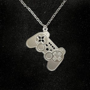 May include: A silver-colored pendant necklace featuring a detailed video game controller design. The controller pendant is suspended from a delicate chain, showcasing buttons and directional pads. The necklace is displayed against a dark background.