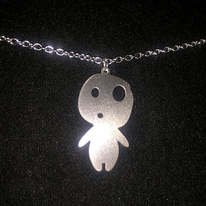 May include: A silver-colored pendant necklace featuring a stylized figure with large, round eyes and small arms. The pendant hangs from a delicate silver chain. The background is a dark, textured surface.