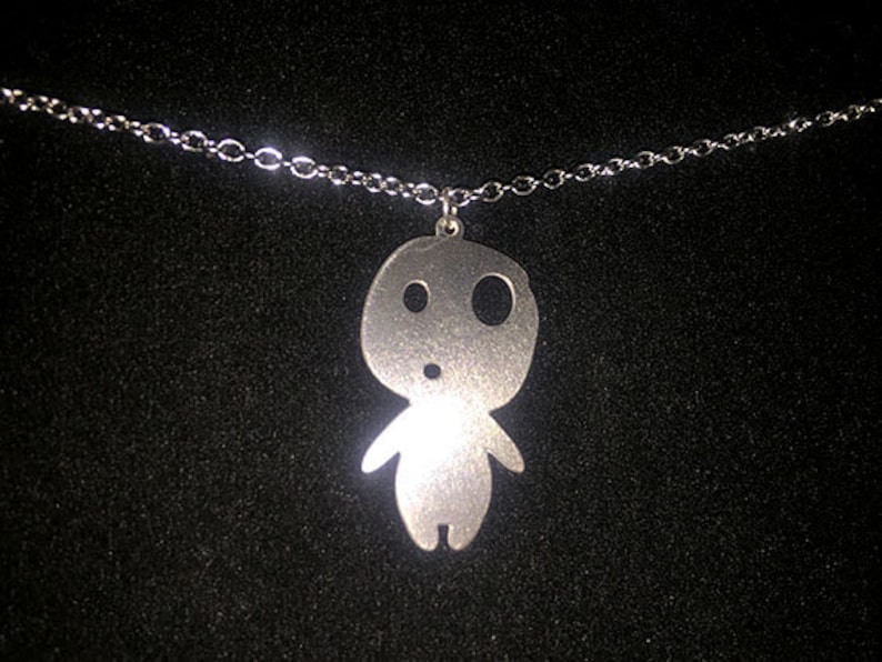 May include: A silver-colored pendant necklace featuring a stylized figure with large, round eyes and small arms. The pendant hangs from a delicate silver chain. The background is a dark, textured surface.