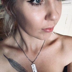 Puede incluir: Un collar de cadena de plata con un colgante rectangular que presenta un dise&ntilde;o de nota musical. El collar lo lleva una persona con un tatuaje de pluma en el hombro.