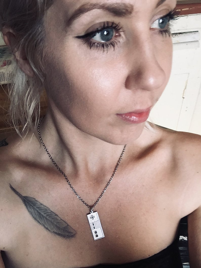 Puede incluir: Un collar de cadena de plata con un colgante rectangular que presenta un dise&ntilde;o de nota musical. El collar lo lleva una persona con un tatuaje de pluma en el hombro.