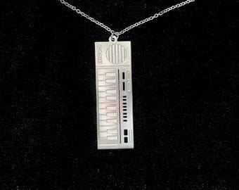 May include: A silver necklace with a pendant shaped like a Casio keyboard. The pendant has the word "CASIO" printed on it.