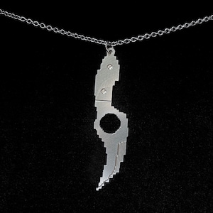 Könnte beinhalten: Eine silberfarbene Halskette mit einem pixeligen Messeranhänger. Das Messer hat ein kreisförmiges Loch und kleine, funkelnde Verzierungen. Die Halskette hängt an einer feinen Silberkette vor schwarzem Hintergrund.
