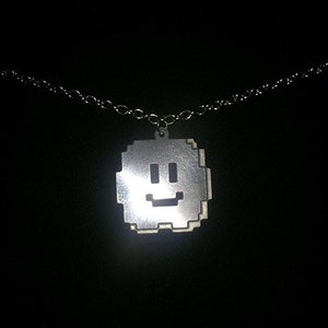 Op de afbeelding: Zilverkleurige ketting met een pixelachtig smiley-gezicht. De vierkante hanger heeft een eenvoudig, vrolijk gezicht. De fijne zilveren ketting contrasteert mooi tegen de donkere achtergrond.