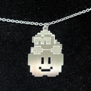 Op de afbeelding: Een zilveren ketting met een gepixelde karakter hanger. Het karakter heeft een glimlachend gezicht en een kapsel.