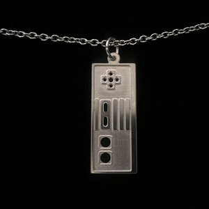 Puede incluir: Un collar de plata con un colgante con forma de mando de videojuego vintage. El colgante tiene un botón en forma de cruz, cuatro botones redondos y un botón rectangular.