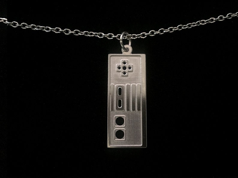 Puede incluir: Un collar de plata con un colgante con forma de mando de videojuego vintage. El colgante tiene un bot&oacute;n en forma de cruz, cuatro botones redondos y un bot&oacute;n rectangular.
