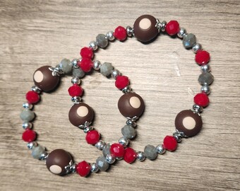 May include: Two beaded bracelets. Each bracelet features alternating red, gray, and silver beads, with larger brown beads that have a white circle in the center. The bracelets are displayed on a wooden surface.