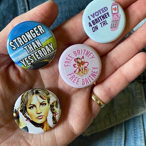 May include: Four button pins with different designs. One pin features a blue background with the text "Stronger Than Yesterday". Another pin features a pink background with the text "Free Britney" and a cartoon image of Britney Spears. A third pin features a blue background with the text "I Voted 4 Britney On Trial" and a cartoon image of a pink cell phone. The fourth pin features a photo of Britney Spears.