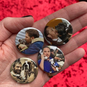 May include: Four round button pins featuring a man with a dog. Each pin features a different photo of the man and dog. The man is wearing a variety of clothing, including a blue shirt, a black jacket, and a gray shirt. The dogs are all different breeds, including a black and white dog, a brown and white dog, and a white dog.