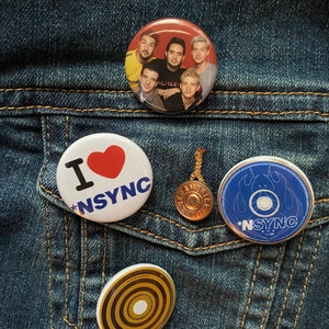 May include: A collection of *NSYNC themed buttons and a pendant on a denim jacket. Buttons include a group photo, the words "I ❤️ *NSYNC", and a blue button with the band's name. The pendant is circular and gold-toned.