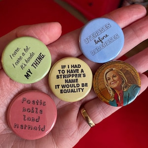 May include: Five round button pins with different sayings. The pins are held in a hand. The pins are light blue, yellow, green, pink, and have a picture of a woman on them. The sayings on the pins are "UTERUSES before DUDERUSES", "IF I HAD TO HAVE A STRIPPER'S NAME IT WOULD BE EQUALITY", "I care. I care a lot. It's kinda MY THING.", "Poetic noble land Mermaid", and "UTERUSES before DUDERUSES".