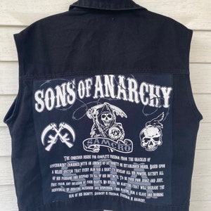 May include: Black denim vest with a large white patch on the back that says "Sons of Anarchy" with a skull and crossbones design. The patch also includes the text "SAMCRO" and a quote about freedom and anarchy.