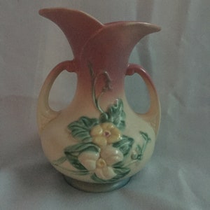 Pode incluir: Um vaso de cerâmica vintage com um esmalte rosa e branco. O vaso tem duas alças e é decorado com flores amarelas e brancas e folhas verdes.
