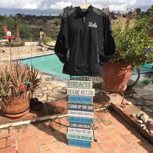 Puede incluir: Una chaqueta negra y verde con la palabra "Ucla" bordada en la parte delantera. La chaqueta est&aacute; colgada en un estante frente a una piscina. Hay ocho letreros de madera apilados uno encima del otro que dicen "Reglas de la casa de playa" y enumeran ocho reglas para disfrutar de la playa.