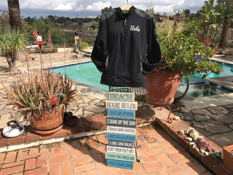 Puede incluir: Una chaqueta negra y verde con la palabra "Ucla" bordada en la parte delantera. La chaqueta est&aacute; colgada en un estante frente a una piscina. Hay ocho letreros de madera apilados uno encima del otro que dicen "Reglas de la casa de playa" y enumeran ocho reglas para disfrutar de la playa.