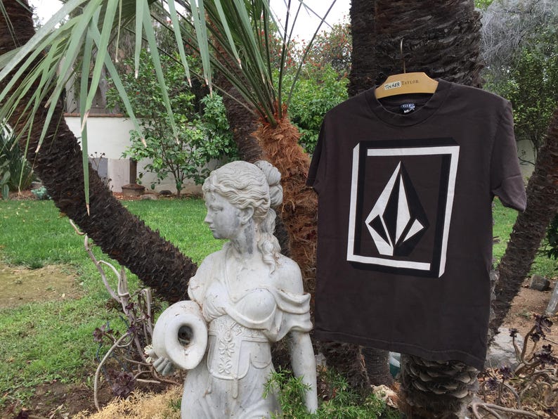 Pu&ograve; includere: Una maglietta marrone scuro con un logo bianco a forma di diamante con un diamante pi&ugrave; piccolo all'interno. Il logo &egrave; all'interno di un quadrato bianco con un bordo nero. La maglietta &egrave; appesa a un ramo d'albero.