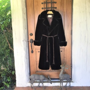 May include: A long, black faux fur coat with a matching belt hangs on a wooden door. The coat features a wide collar, cuffs, and a brown braided belt. The door is brown with a metal handle. Two deer statues stand in front of the door.
