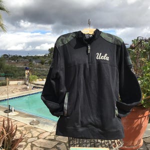 Puede incluir: Sudadera negra de cremallera de un cuarto con mangas de camuflaje y un logotipo "UCLA" blanco en la parte delantera.