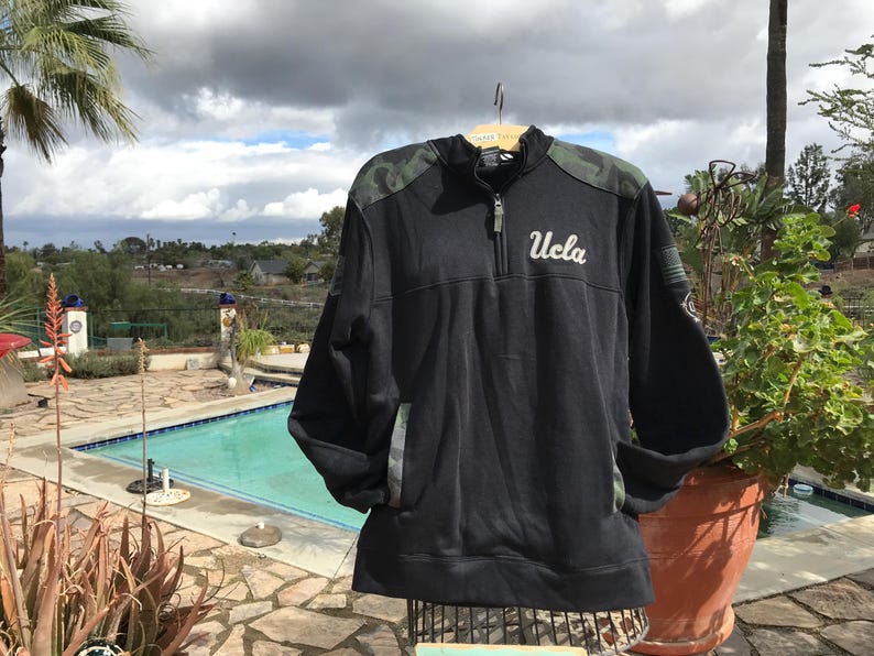 Puede incluir: Sudadera negra de cremallera de un cuarto con mangas de camuflaje y un logotipo "UCLA" blanco en la parte delantera.