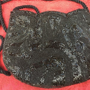 May include: A black beaded evening bag with a rope strap. The bag is decorated with a swirling pattern of black beads.