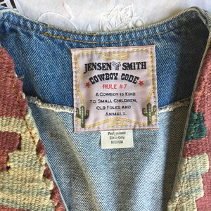 K&ouml;nnte beinhalten: Eine Jeansweste mit einem gewebten Wandteppichmuster auf Vorder- und R&uuml;ckseite. Die Weste hat ein Etikett mit der Aufschrift "Jensen Smith Cowboy Code Rule #7 A cowboy is kind to small children, old folks and animals. Professional Clean Only Medium."