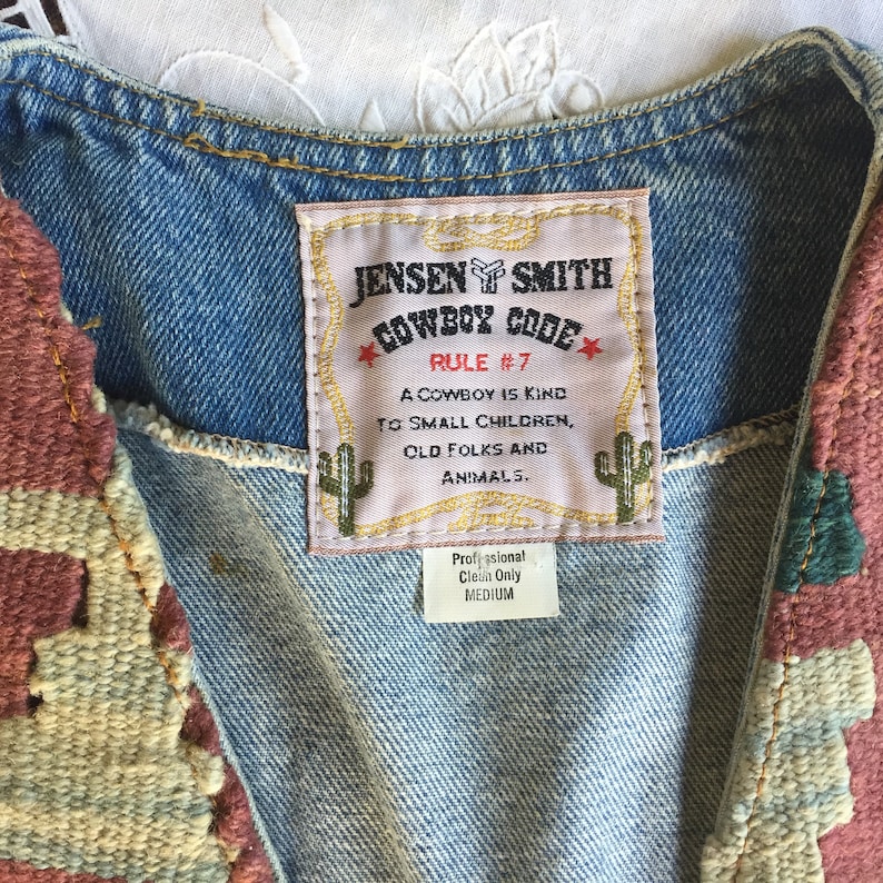 K&ouml;nnte beinhalten: Eine Jeansweste mit einem gewebten Wandteppichmuster auf Vorder- und R&uuml;ckseite. Die Weste hat ein Etikett mit der Aufschrift "Jensen Smith Cowboy Code Rule #7 A cowboy is kind to small children, old folks and animals. Professional Clean Only Medium."