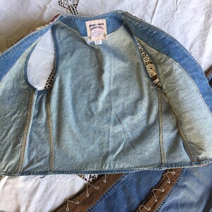 K&ouml;nnte beinhalten: Eine Jeansweste mit einem braunen und wei&szlig;en Blumenmuster auf der R&uuml;ckseite. Die Weste ist offen und das Innere ist sichtbar. Die Weste hat ein Etikett mit der Aufschrift "Johnny & Smith Country Style".