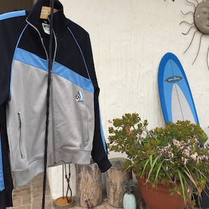 May include: A black, gray, and light blue zip-up track jacket with a high collar. The jacket features a diagonal color block design and a small logo on the chest. The sleeves have light blue stripes. The jacket is hanging on a wooden hanger.