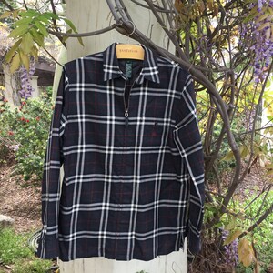 May include: A black and white plaid long-sleeve shirt with a red zipper and a small pocket on the left chest. The shirt is hanging on a wooden hanger.