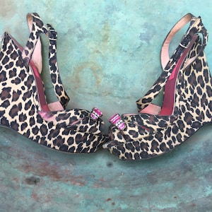 May include: A pair of leopard print wedge sandals with pink and clear rhinestone embellishments. The sandals have a closed toe and an ankle strap.