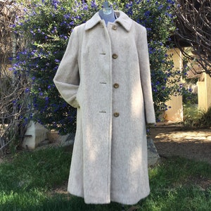 May include: A long, light beige wool coat with a collar and button closure. The coat has a slightly flared shape and is shown on a mannequin in a garden setting.