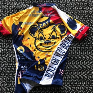 May include: A yellow, red, and navy blue cycling jersey with a cartoon bear graphic. The jersey has the text "ROLL ON YOU BEARS!" and "545 miles - 7 days San Francisco to Los Angeles". The jersey is made of a breathable material.