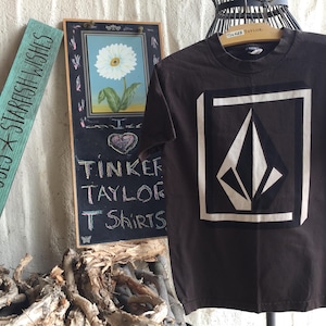 May include: A brown t-shirt with a white diamond logo and the text "Tinker Taylor T Shirts" on a chalkboard in the background.